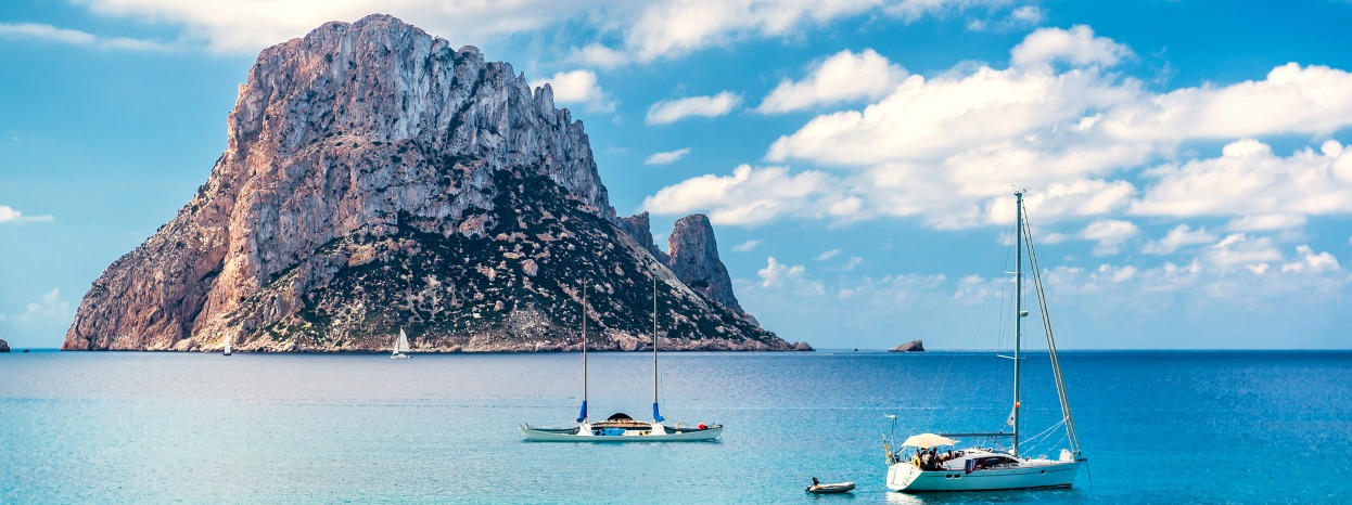 Boats sailing in a turquoise sea, a giant rock formation is seen in the background.