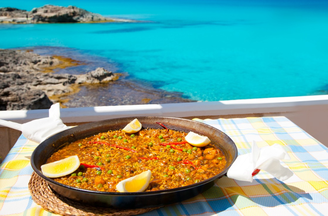 A pan with paella on a table by the ocean.