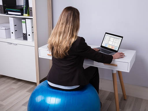Woman sits on exercise ball while working at desk on laptop.