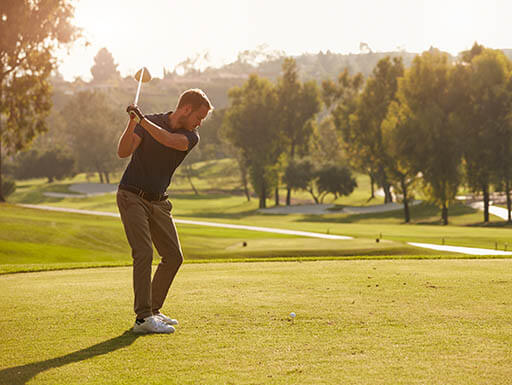 Man in a dark polo and khakis swings club on Simi Hills Golf Course in Simi Valley, CA, on sunny morning with fairway, cart path and leafy green trees in the background Man in a dark polo and khakis swings club on Simi Hills Golf Course in Simi Valley, CA, on sunny morning with fairway, cart path and leafy green trees in the background