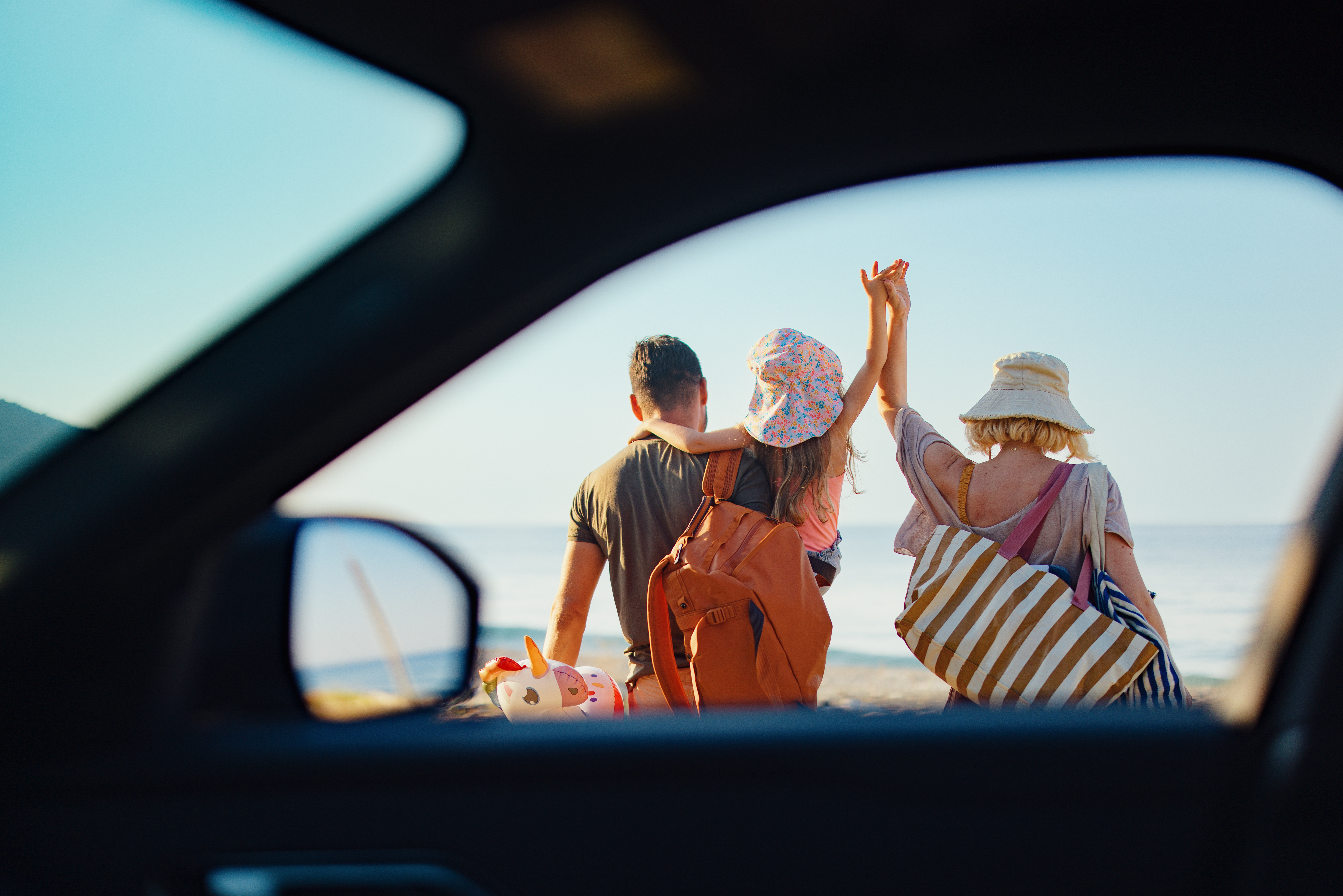 2214123145 A family enjoys a seaside outing, captured through a unique vehicle frame perspective. With smiles, accessories, and ocean waves, this image encapsulates joyful summer vacations and togetherness by the beach under clear skies.